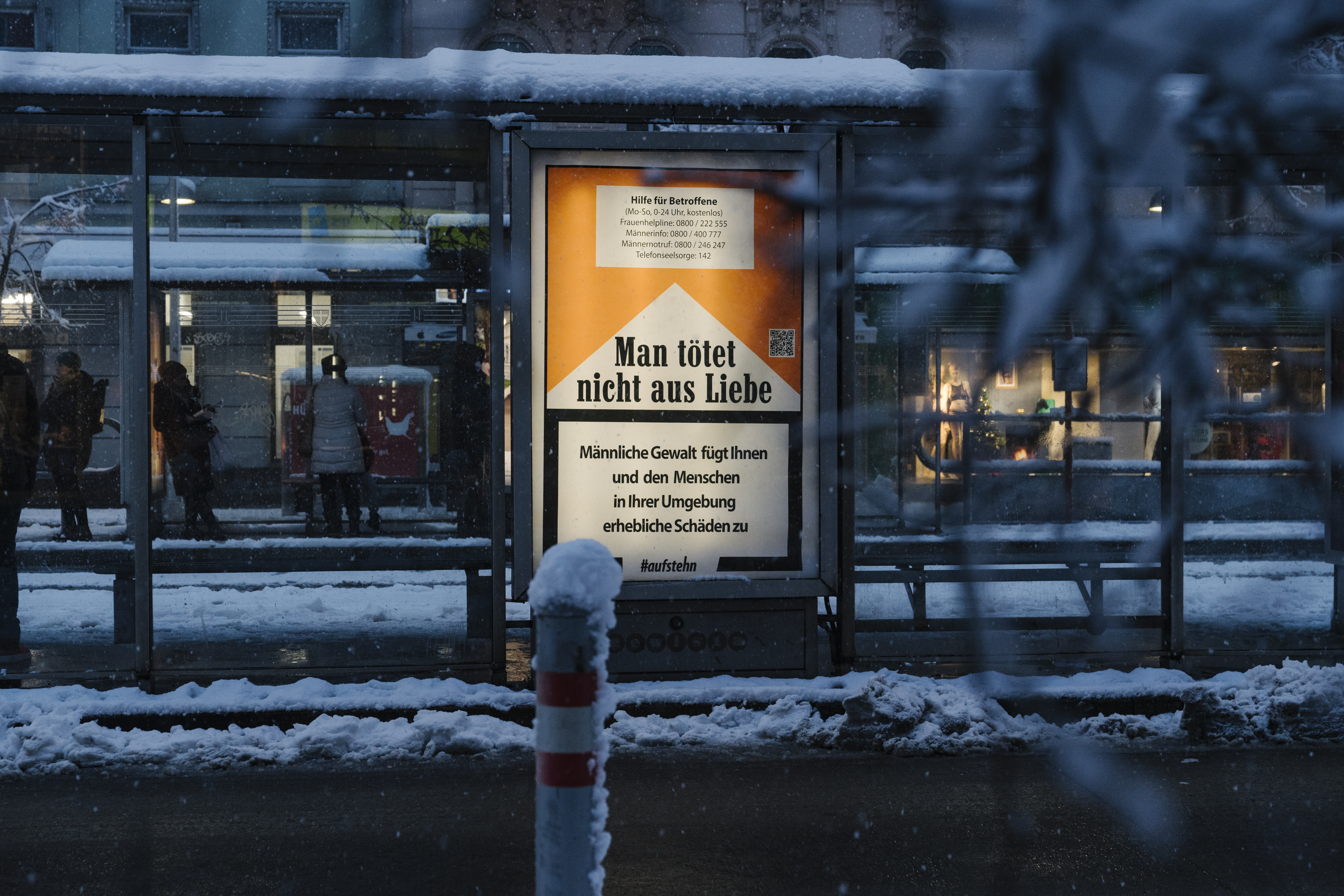 Plakat mit Aufschrift "Man tötet nicht aus Liebe - Männliche Gewalt fügt Ihnen und den Menschen in Ihrer Umgebung erhebliche Schäden zu" im Stil der Warnungen auf Zigarettenschachteln an einer verschneiten Bushaltestelle.
