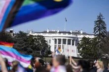 Das Weiße Haus, Präsidentschaftssitz der USA, ist leicht schräg im Fokus der Betrachtenden. Davor, in leichter Unschärfe zeichnet sich ein Demonstrationszug mit LBGTQIA+ Flaggen ab.