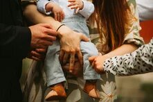 Nodoption Ein Baby wird von zwei weißen weiblichen Händen gehalten. Eine weitere Hand nähert sich fürsorglich dem Bein des Kindes. Ein weiteres Händepaar ist ganz in seiner Nähe. Die Szene wirkt wohlig, kümmernd, dynamisch.