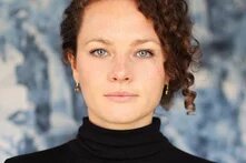 Chiara Fleischhacker, VENA Regisseurin Eine weiblich gelesene Person sieht mit blaugrauen Augen direkt die betrachtende Person an. Sie hat braune gelockte Haare, die sie hochgesteckt trägt. Der Gesichtsausdruck ist ruhig und selbstsicher.
