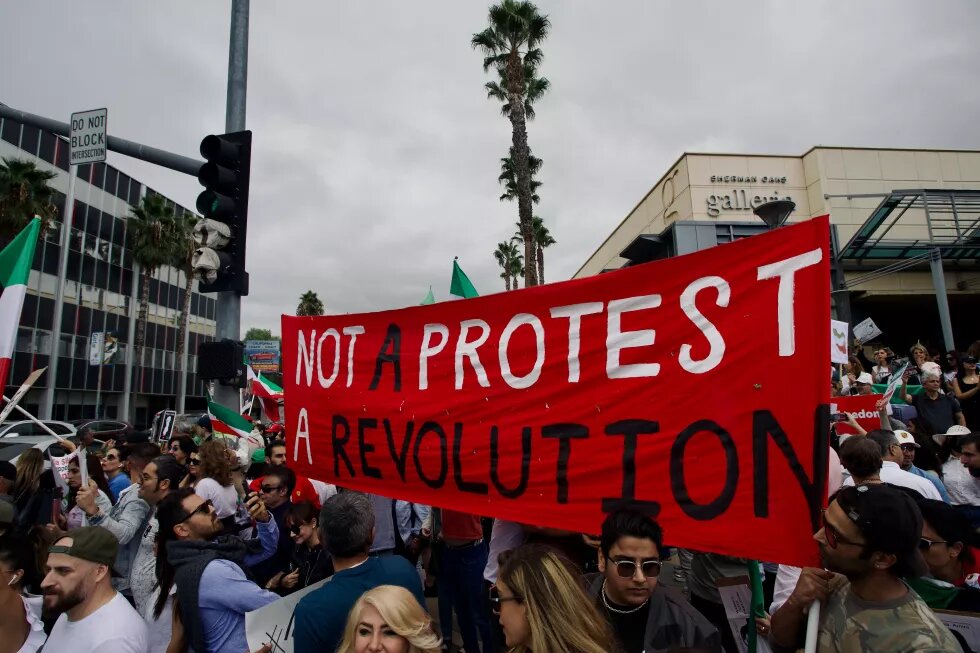 Auf em Foto ist eine Demonstration zu sehen, mehrere iranische Flaggen werden hochgehalten. In der Mitte ist ein rotes Banner, auf dem Steht - Not a protest, a revolution. Im linken oberen Bildrand ist ein Straßenschild abgebildet, auf dem steht: Do not block intersection. 