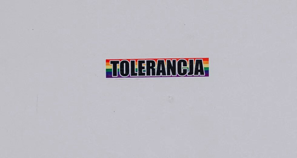 Auf einem Sticker mit Regenbogen Hintergrund steht auf polnisch das Wort "Toleranz". Der Sticker klebt auf einer weißen Wand.