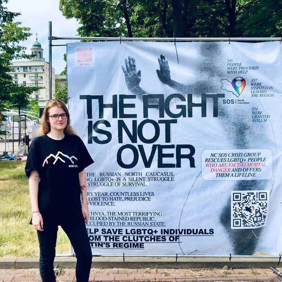 Eine Person mit langen dunklen Haaren und schwarzer Kleidung steht links neben einem Plakat und gehört der Gruppe "North Caucasus Crisis Group" an. Die Gruppe ist bei der Aufnahme auf der Pride in Warschau am 17. Juni 2023.