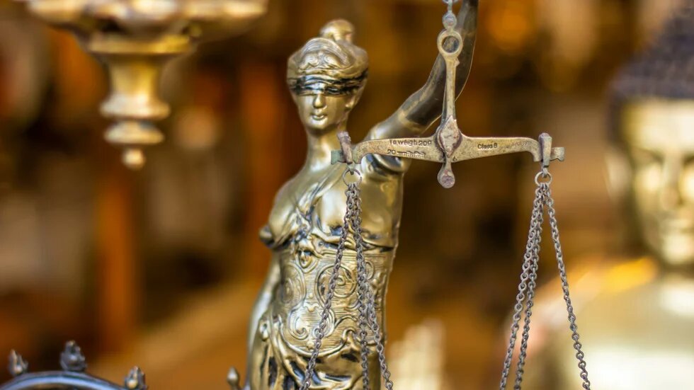 Es wird eine Justicia-Figur in goldener Farbe im Anschnitt dargstellt. Der Hintergrund ist verschwommen.