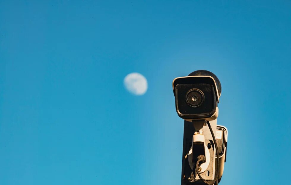 Blauer Himmer, Mond in Tiefenunschärfe, davor eine Kamera auf einem Mast staffiert die die Betrachtenden fokussiert