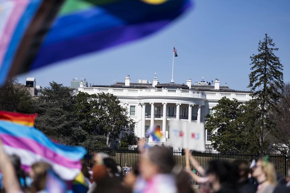 Das Weiße Haus, Präsidentschaftssitz der USA, ist leicht schräg im Fokus der Betrachtenden. Davor, in leichter Unschärfe zeichnet sich ein Demonstrationszug mit LBGTQIA+ Flaggen ab.