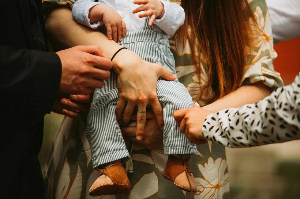 Nodoption Ein Baby wird von zwei weißen weiblichen Händen gehalten. Eine weitere Hand nähert sich fürsorglich dem Bein des Kindes. Ein weiteres Händepaar ist ganz in seiner Nähe. Die Szene wirkt wohlig, kümmernd, dynamisch.