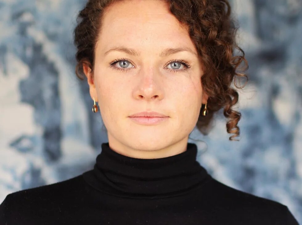 Chiara Fleischhacker, VENA Regisseurin Eine weiblich gelesene Person sieht mit blaugrauen Augen direkt die betrachtende Person an. Sie hat braune gelockte Haare, die sie hochgesteckt trägt. Der Gesichtsausdruck ist ruhig und selbstsicher.