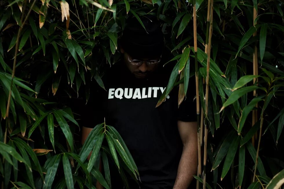 Eine Person steht vor Pflanzen. Auf ihrem T-Shirt steht: Equality