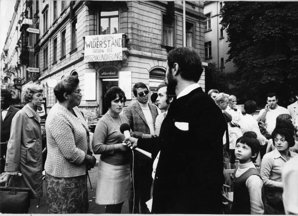 Eine Gruppe Menschen steht vor einem Haus mit einem Banner "Widerstand gegen Massenkündigungen" 