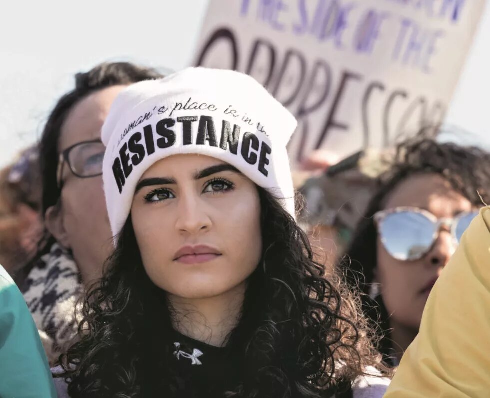 Eine Frau mit einer Mütze steht in einer Demonstration. Auf ihrer Mütze ist gestickt: Women's place is in the resistance