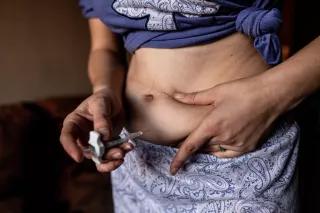 Auf dem Bild sieht man eine Person in Rock und hochgewickeltem Tshirt, die sich gerade selbst eine Spritze in den Bauch gibt.